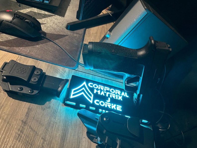 May include: A black handgun, holster, and magazine are displayed on a wooden surface. A glowing blue sign reads "CORPORAL MATRIX J CORKE" with a military rank insignia. A computer mouse and speaker are also visible.