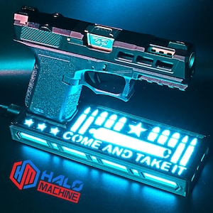 May include: Black and copper-colored handgun with a textured grip, resting above a glowing rectangular base. The base features the text "COME AND TAKE IT" and bullet illustrations, with the Halo Machine logo visible below.