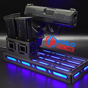 May include: A black handgun rests on a black base with blue LED lighting. Two black magazines are in a holder to the left. The base has the text "HALO MACHINE" in red and blue.