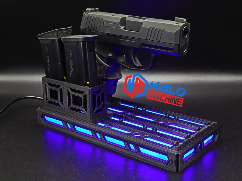 May include: A black handgun rests on a black base with blue LED lighting. Two black magazines are in a holder to the left. The base has the text "HALO MACHINE" in red and blue.
