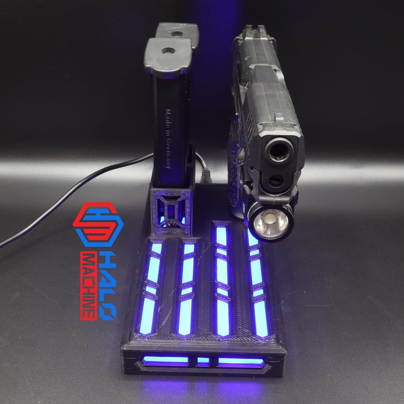 May include: A black gun display stand with blue LED lights. The stand holds a black handgun with a mounted flashlight. The stand has the text "Made in Germany" and the logo "HALO MACHINE".