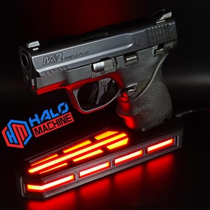 May include: A black handgun with the text "M&P 9 SHIELD PLUS" rests above a rectangular base with red glowing lights. The base has the text "HALO MACHINE" in red and blue. The background is black.