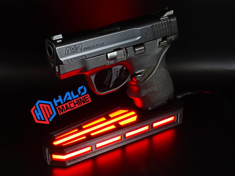 May include: A black handgun with the text "M&P 9 SHIELD PLUS" rests above a rectangular base with red glowing lights. The base has the text "HALO MACHINE" in red and blue. The background is black.