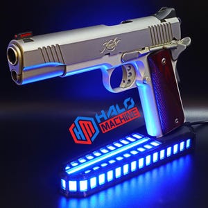 May include: A silver handgun with a wooden grip, resting on a black base with blue LED lights. The text "HALO MACHINE" is visible on the base. The gun has a red sight and the brand name "Kimber" is engraved on the slide.
