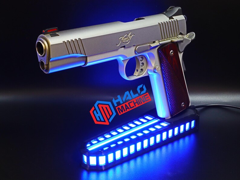 May include: A silver handgun with a wooden grip, resting on a black base with blue LED lights. The text "HALO MACHINE" is visible on the base. The gun has a red sight and the brand name "Kimber" is engraved on the slide.