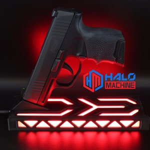 May include: Black handgun resting on a black base with red LED lighting. The base has a geometric design and the text "HALO MACHINE" is visible. The image is set against a dark background.