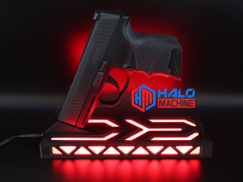 May include: Black handgun resting on a black base with red LED lighting. The base has a geometric design and the text "HALO MACHINE" is visible. The image is set against a dark background.