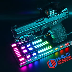 May include: A black handgun with a red dot sight and a mounted light, resting on a base with colorful LED lights. The base has the text "HALO MACHINE" and a logo. The gun is a HK VP9.