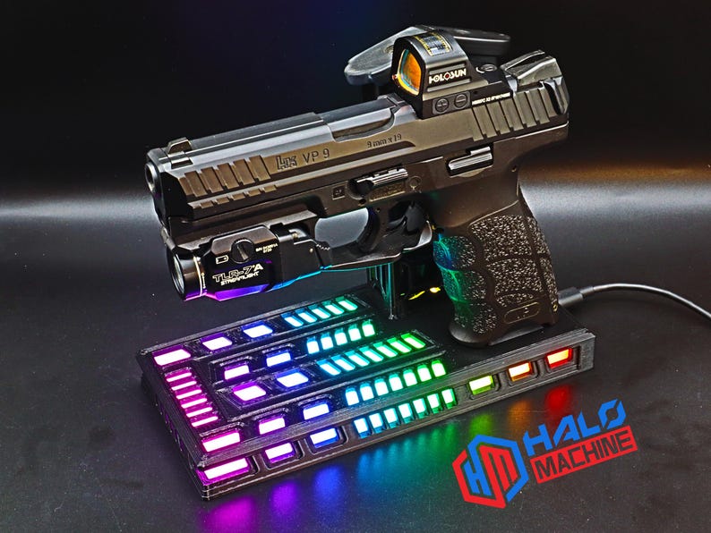 May include: A black handgun with a red dot sight and a mounted flashlight rests on a black base with a colorful LED light display. The base has the text "HALO MACHINE" in red and blue.