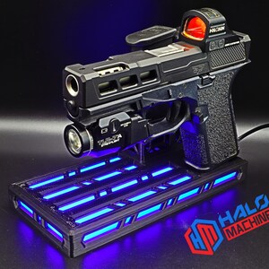 May include: Black handgun with a red dot sight and a mounted flashlight, resting on a black base with blue LED lights. The base has the text "HALO MACHINE" in red and blue.