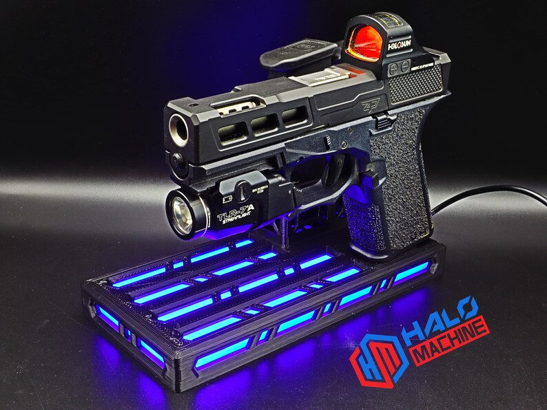 May include: Black handgun with a red dot sight and a mounted flashlight, resting on a black base with blue LED lights. The base has the text "HALO MACHINE" in red and blue.