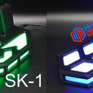 May include: Two black electronic devices with illuminated LED lights. One glows green, labelled "SK-1", and the other glows blue, labelled "SK-2". Both feature the "HALO MACHINE" logo. The devices have a geometric design and a rectangular base.