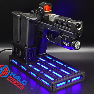 May include: Black handgun with a red dot sight and a mounted flashlight, resting on a black base with blue LED lights. Two black magazines are positioned to the left. The base has the text "HALO MACHINE" in red and blue.