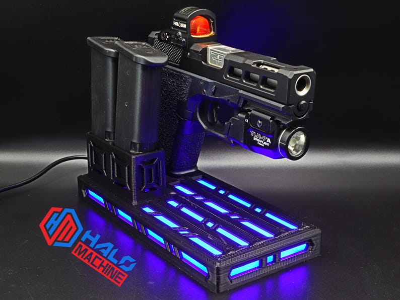 May include: Black handgun with a red dot sight and a mounted flashlight, resting on a black base with blue LED lights. Two black magazines are positioned to the left. The base has the text "HALO MACHINE" in red and blue.