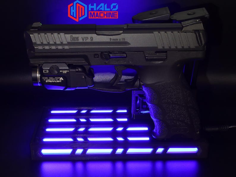 May include: A black handgun with the text "HK VP 9" and "9 mm x 19" is displayed on a stand with blue LED lights. A TLR-7A Streamlight is attached to the gun. The Halo Machine logo is visible in the background.