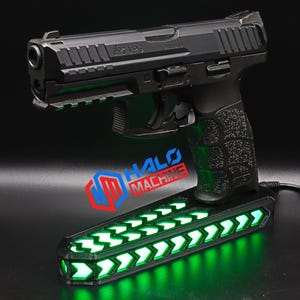 May include: A black handgun with a textured grip rests on a base with green LED lights. The base has a series of illuminated arrows. The text "HALO MACHINE" is visible on the base.