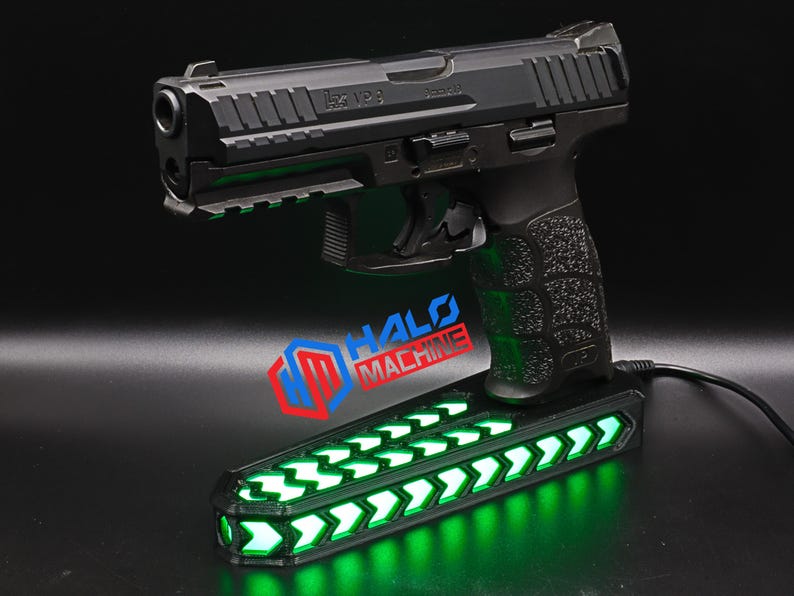 May include: A black handgun with a textured grip rests on a base with green LED lights. The base has a series of illuminated arrows. The text "HALO MACHINE" is visible on the base.