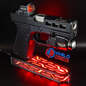 May include: A black handgun with a red dot sight and a mounted flashlight. The gun is resting on a red-lit base with the text "HALO MACHINE". The image is set against a dark background.