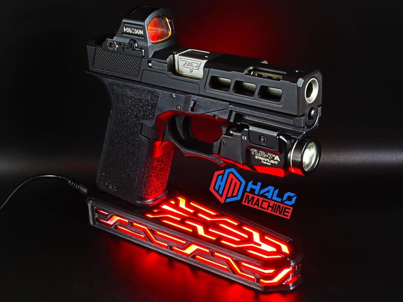 May include: A black handgun with a red dot sight and a mounted flashlight. The gun is resting on a red-lit base with the text "HALO MACHINE". The image is set against a dark background.