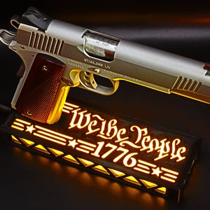 May include: A silver and gold handgun with a red grip rests above a black rectangular light box. The light box displays the words "We the People 1776" in a glowing, stylized font, with star and stripe accents. The overall theme is patriotic.