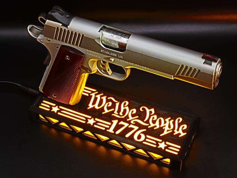 May include: A silver and gold handgun with a red grip rests above a black rectangular light box. The light box displays the words "We the People 1776" in a glowing, stylized font, with star and stripe accents. The overall theme is patriotic.