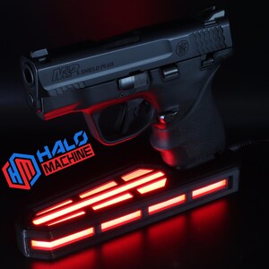 May include: A black handgun, model M&P Shield Plus, rests above a rectangular base with red LED lights. The base has the text "HALO MACHINE" and a logo. The image is set against a dark background.