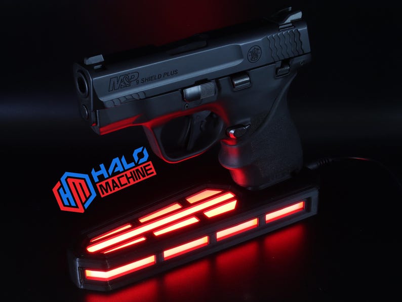 May include: A black handgun, model M&P Shield Plus, rests above a rectangular base with red LED lights. The base has the text "HALO MACHINE" and a logo. The image is set against a dark background.