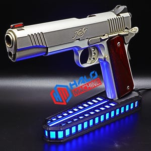 May include: A silver handgun with a wooden grip rests above a black base with blue LED lights. The handgun has a red sight and the brand name "Kimber" is visible. The base has the text "HALO MACHINE".