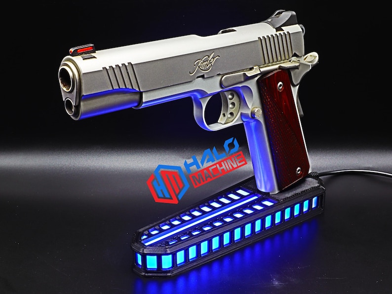 May include: A silver handgun with a wooden grip rests above a black base with blue LED lights. The handgun has a red sight and the brand name "Kimber" is visible. The base has the text "HALO MACHINE".
