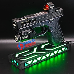 Puede incluir: Una pistola negra con una mira de punto rojo y una luz verde en un soporte negro con luces verdes. El soporte está conectado a una fuente de alimentación mediante un cable negro.
