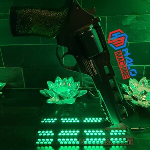 May include: A black Rhino 60DS revolver is displayed against a green-lit backdrop. The gun is positioned above a rectangular base with numerous green LED lights. The Halo Machine logo is visible. Decorative lotus flower-shaped lights are also present.