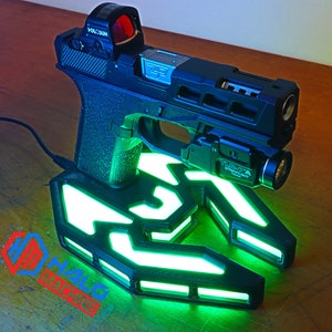 May include: Black handgun with a red dot sight and a mounted flashlight, on a custom charging base. The base has glowing green accents and the text "HALO MACHINE". The gun is connected to a power source.