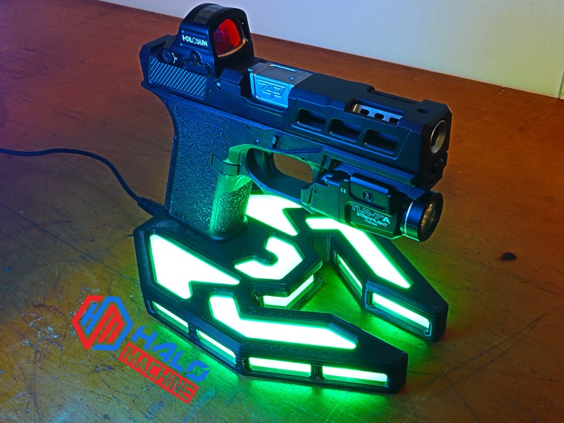 May include: Black handgun with a red dot sight and a mounted flashlight, on a custom charging base. The base has glowing green accents and the text "HALO MACHINE". The gun is connected to a power source.
