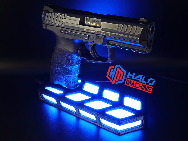 May include: A black handgun is displayed on a platform with bright blue lights. The platform has a geometric design. The words "HALO MACHINE" are in red and blue. The background is dark.