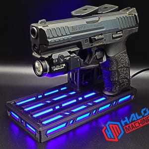 May include: A black handgun with a mounted flashlight rests on a black stand with blue LED lights. The stand has the text "HALO MACHINE" in red and blue. The handgun is a HK VP9.