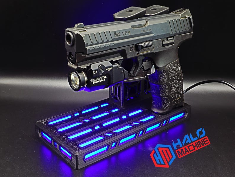 May include: A black handgun with a mounted flashlight rests on a black stand with blue LED lights. The stand has the text "HALO MACHINE" in red and blue. The handgun is a HK VP9.