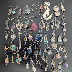 May include: A collection of assorted silver-toned earrings featuring various designs including a cat silhouette, butterfly, heart, cross, turtle, and star shapes, accented with colorful gemstones, abalone shell, clear crystals, and intricate metalwork.