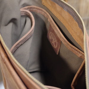 May include: The inside of a brown leather bag with a zipper closure. The bag has a brown fabric lining and a small pocket with the initials "PK" embossed on it.