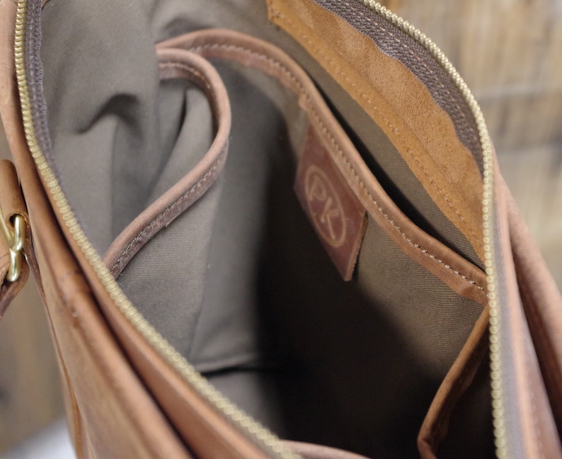 May include: The inside of a brown leather bag with a zipper closure. The bag has a brown fabric lining and a small pocket with the initials "PK" embossed on it.