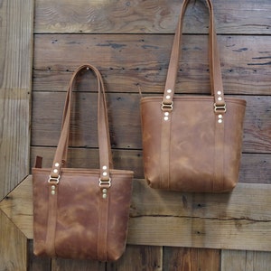 May include: Two brown leather tote bags with brass hardware hanging on a wooden wall. The bags have a distressed look and are different sizes.