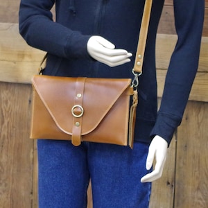 May include: A brown leather crossbody bag with a flap closure and a metal ring detail. The bag has a long adjustable strap and a small tassel.