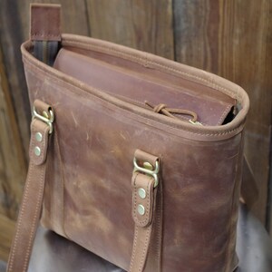May include: A brown leather tote bag with two adjustable straps and brass hardware. The bag has a zipper closure and a brown leather lining.