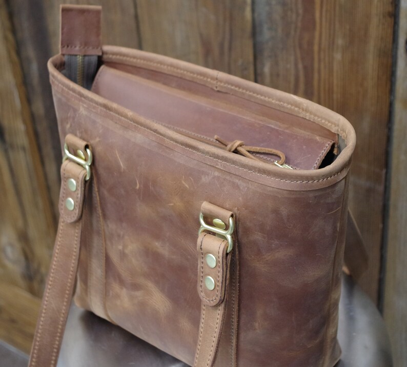May include: A brown leather tote bag with two adjustable straps and brass hardware. The bag has a zipper closure and a brown leather lining.