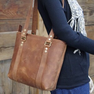 May include: A brown leather tote bag with brass hardware and a top zipper closure. The bag has a wide, flat base and two top handles.