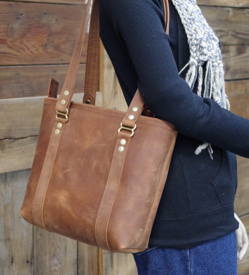 May include: A brown leather tote bag with brass hardware and a top zipper closure. The bag has a wide, flat base and two top handles.