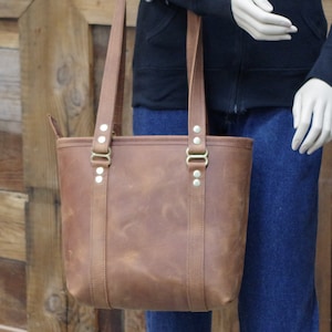 May include: A brown leather tote bag with brass hardware and a top zipper closure. The bag has two long straps and a wide base.