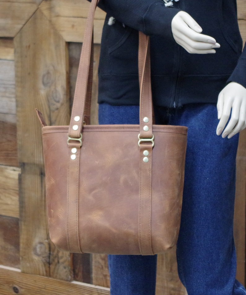 May include: A brown leather tote bag with brass hardware and a top zipper closure. The bag has two long straps and a wide base.
