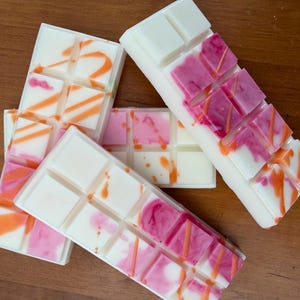 May include: A collection of white wax melt bars with pink and orange detailing, arranged on a wooden surface. The bars are segmented into rectangles, with vibrant pink and orange drizzles and splashes. The overall appearance is bright and appealing.