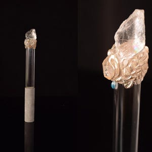 Puede incluir: Un tap&oacute;n de botella de vidrio transparente con una parte superior de cristal decorativa de color claro. El cristal est&aacute; montado sobre una base dorada y ornamentada. El tap&oacute;n tiene un tallo de vidrio transparente y una base de hormig&oacute;n gris.