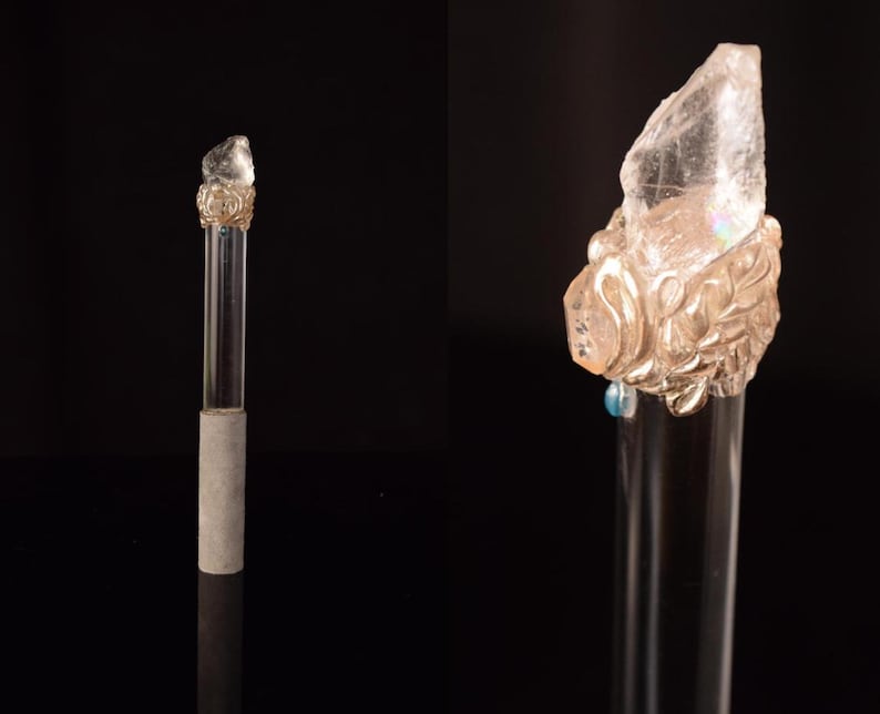 Puede incluir: Un tap&oacute;n de botella de vidrio transparente con una parte superior de cristal decorativa de color claro. El cristal est&aacute; montado sobre una base dorada y ornamentada. El tap&oacute;n tiene un tallo de vidrio transparente y una base de hormig&oacute;n gris.